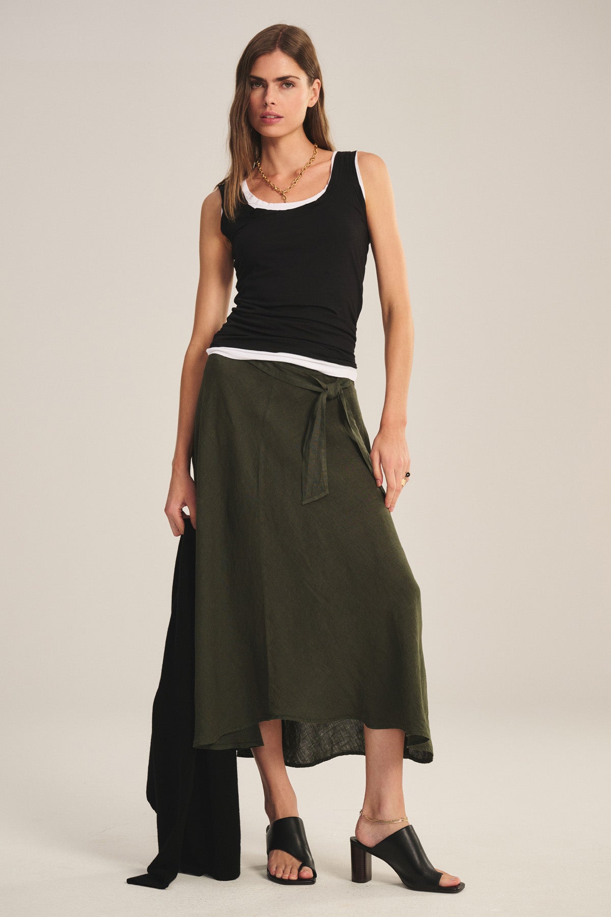 Against a plain background, a woman wears the MALINA LINEN SKIRT by Velvet by Graham & Spencer—an olive green flowy skirt with a high-low hem—paired with black and white layered tank tops and black heeled sandals.