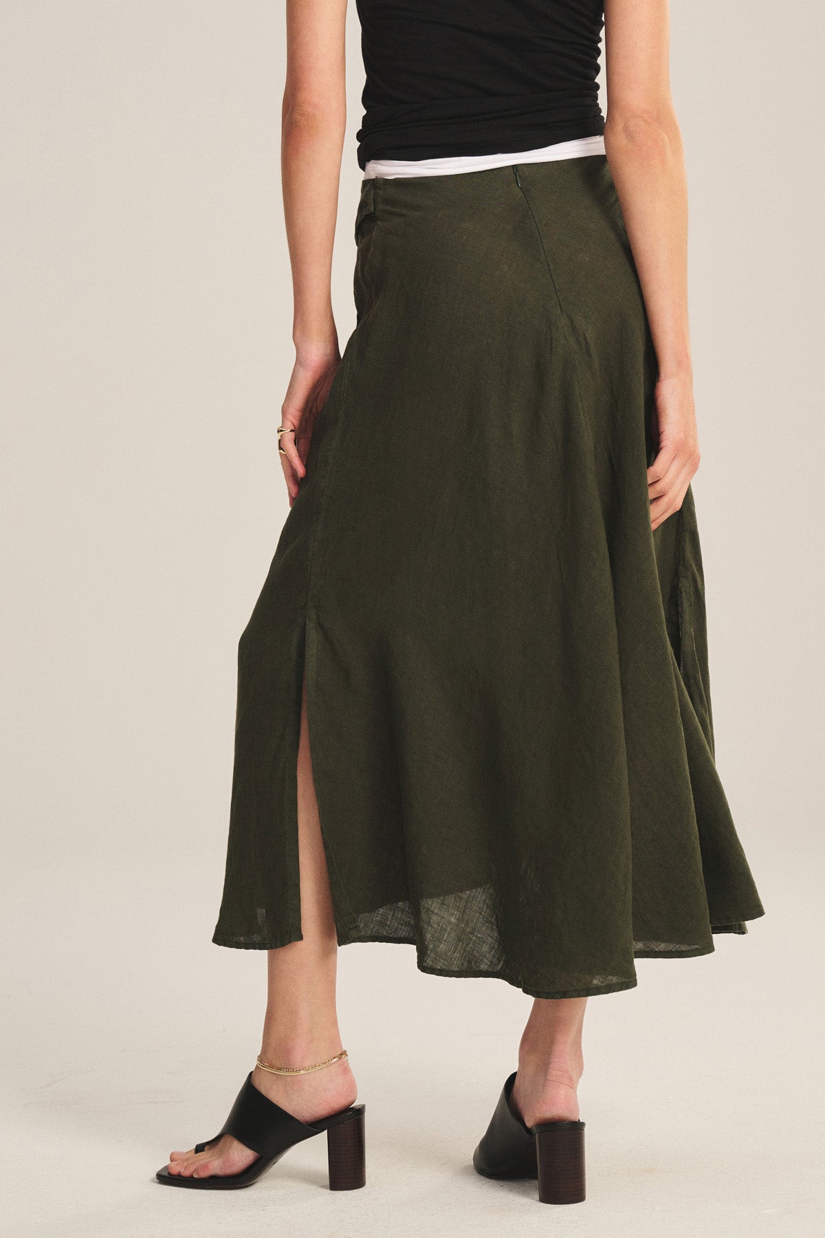 A person stands facing away, wearing a black sleeveless top, black heeled sandals, and the MALINA LINEN SKIRT by Velvet by Graham & Spencer—an olive green flowy skirt with a side slit.-40808415002817