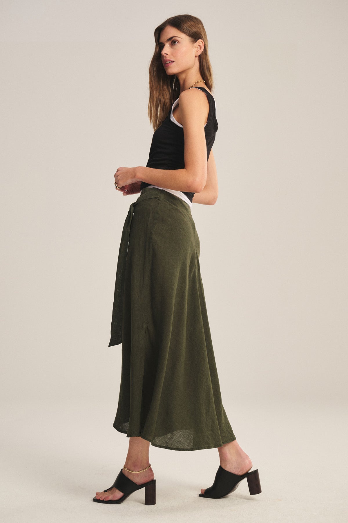 A woman stands sideways against a plain background wearing a black tank top, black heeled mules, and the MALINA LINEN SKIRT by Velvet by Graham & Spencer in olive green with a flowy silhouette and tie waist.-40808414970049