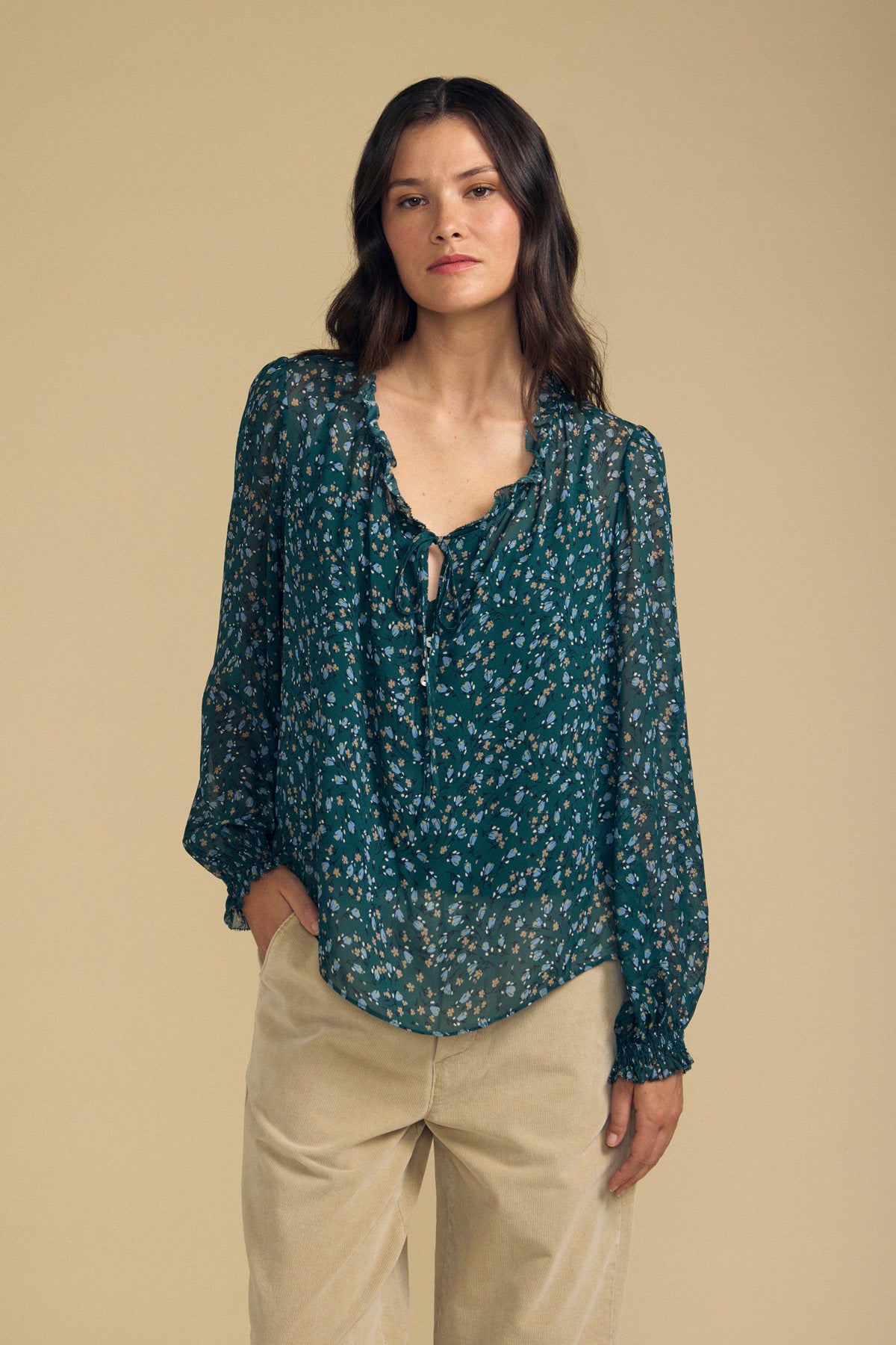Clad in the MARA BLOUSE by Velvet by Graham & Spencer—a sheer, teal floral top made from lightweight viscose with a ruffle neckline—and beige pants, the woman stands before a plain beige background.