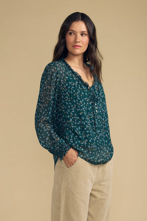 Dressed in the Velvet by Graham & Spencer MARA BLOUSE—a dark green floral top featuring a ruffle neckline—paired with beige pants, the woman stands with her hands in her pockets, gazing to the side against a plain beige background. The lightweight viscose fabric of the blouse brings an effortless elegance to her ensemble.
