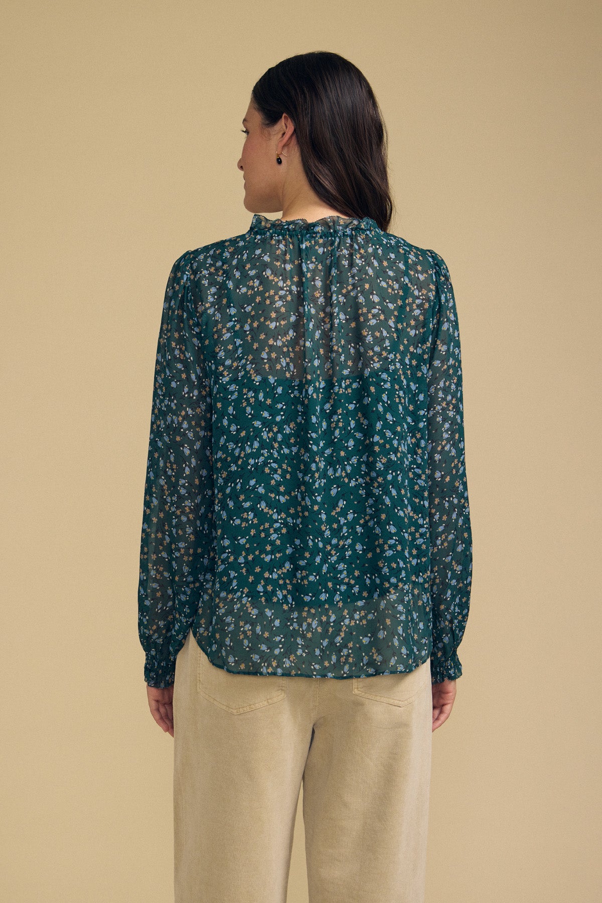 A person stands facing away, wearing the MARA BLOUSE by Velvet by Graham & Spencer—a sheer green floral blouse with a ruffle neckline and long sleeves, crafted from lightweight viscose—paired with beige pants against a plain beige background.-41838221820097