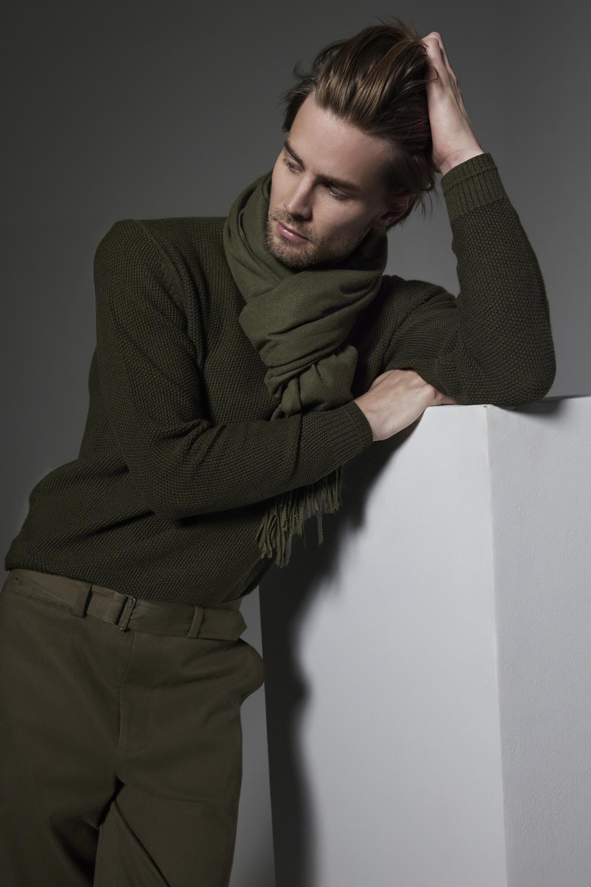   Person wearing the Velvet by Graham & Spencer MARCO SWEATER, featuring a seed stitch texture, along with a scarf and pants. They lean on a white surface with one hand on their head and eyes half-closed, striking a pensive pose against a dark background. 