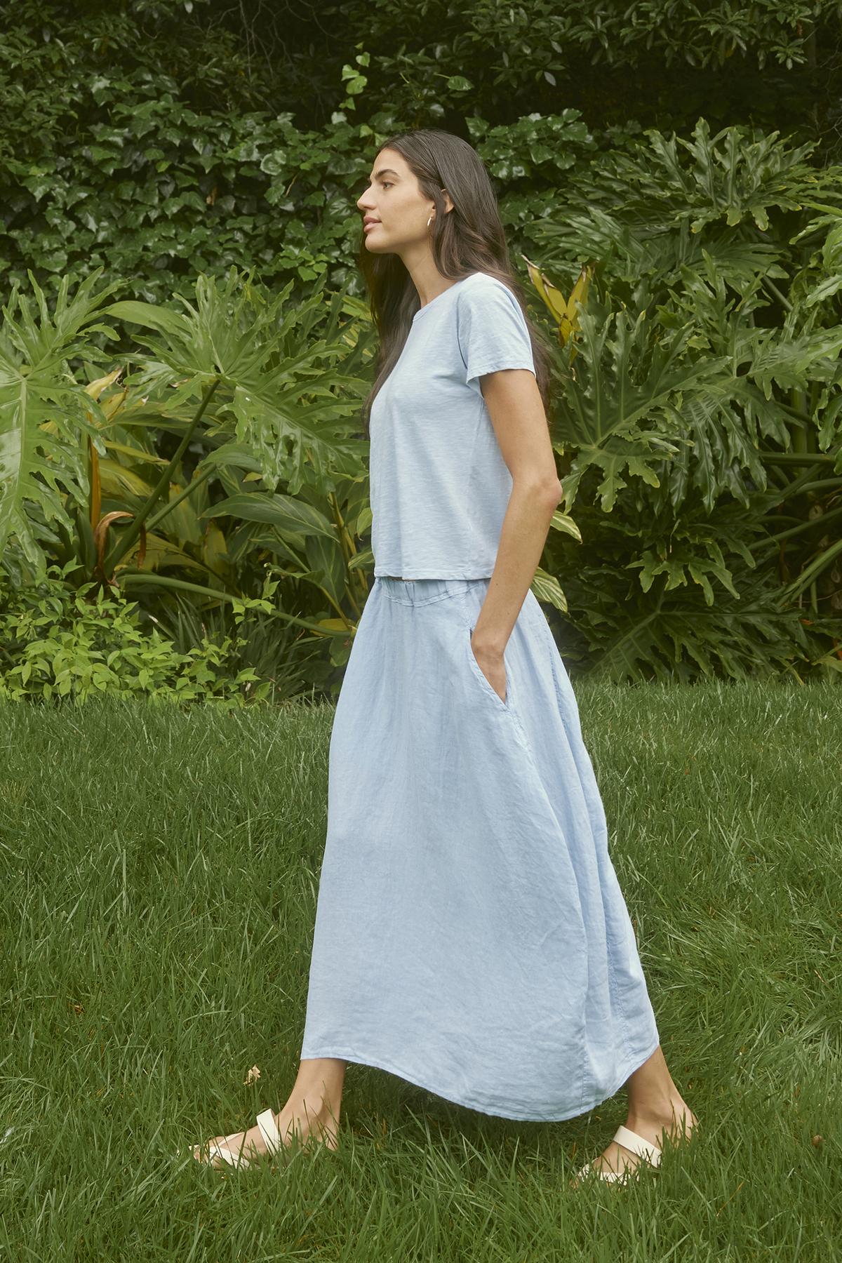 A person with long dark hair wearing a pale blue shirt and a matching FAE LINEN SKIRT by Velvet by Graham & Spencer walks barefoot on grass with lush green foliage in the background.-37104281944257