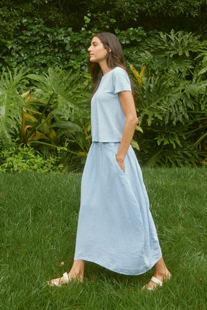 A person with long dark hair wearing a pale blue shirt and a matching FAE LINEN SKIRT by Velvet by Graham & Spencer walks barefoot on grass with lush green foliage in the background.