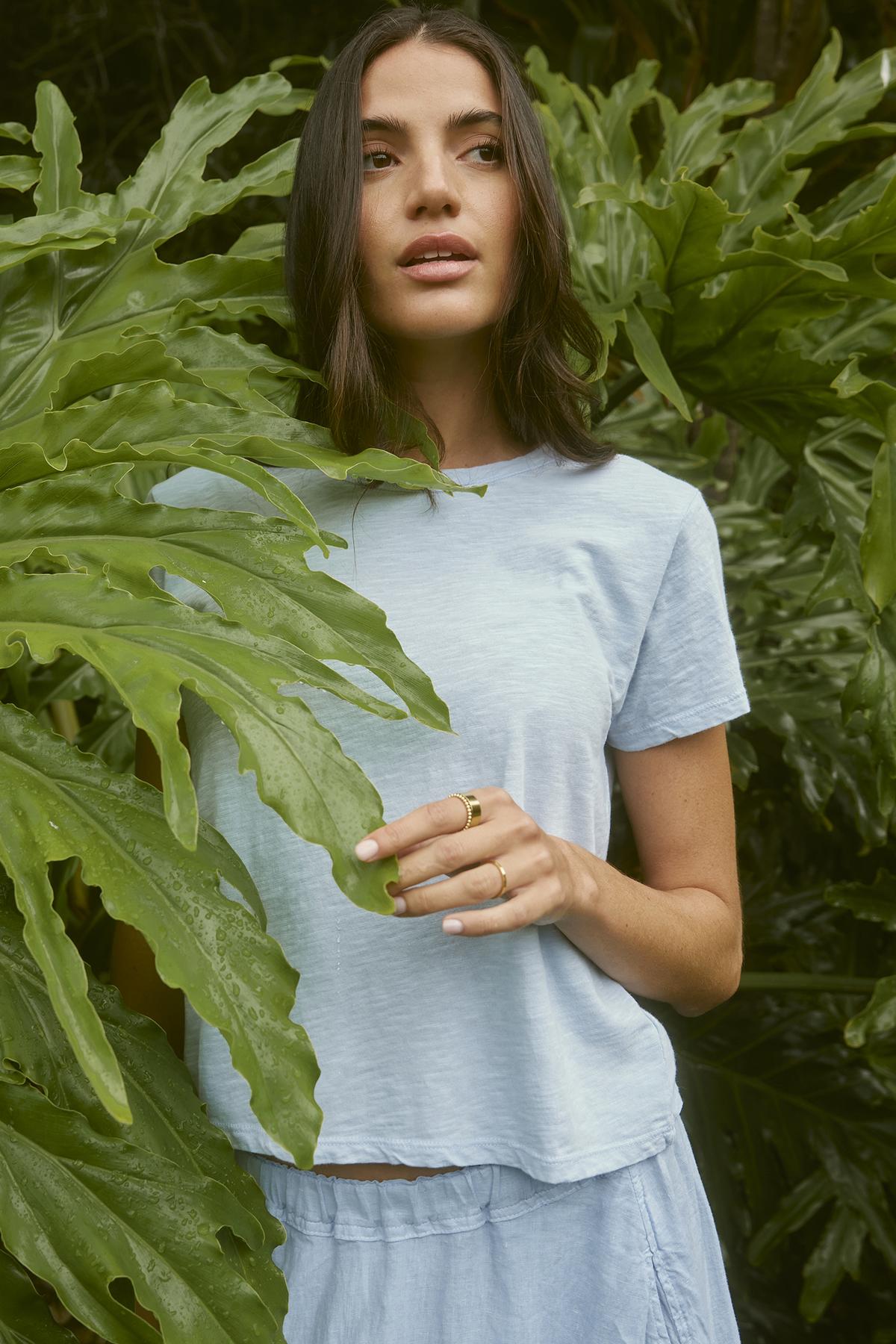   A person with long dark hair stands in a lush green environment, wearing a Velvet by Graham & Spencer MARIE TEE featuring a crew neckline and relaxed boxy fit, while holding a large green leaf. 