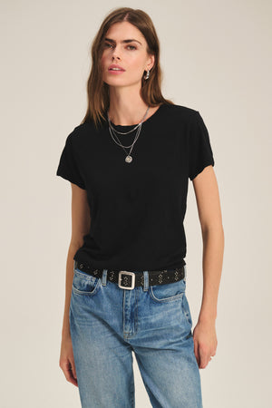A person wearing the boxy fit black MARIKA TEE by Velvet by Graham & Spencer in cotton slub, paired with blue jeans, a studded black belt, and layered silver necklaces stands against a plain background.