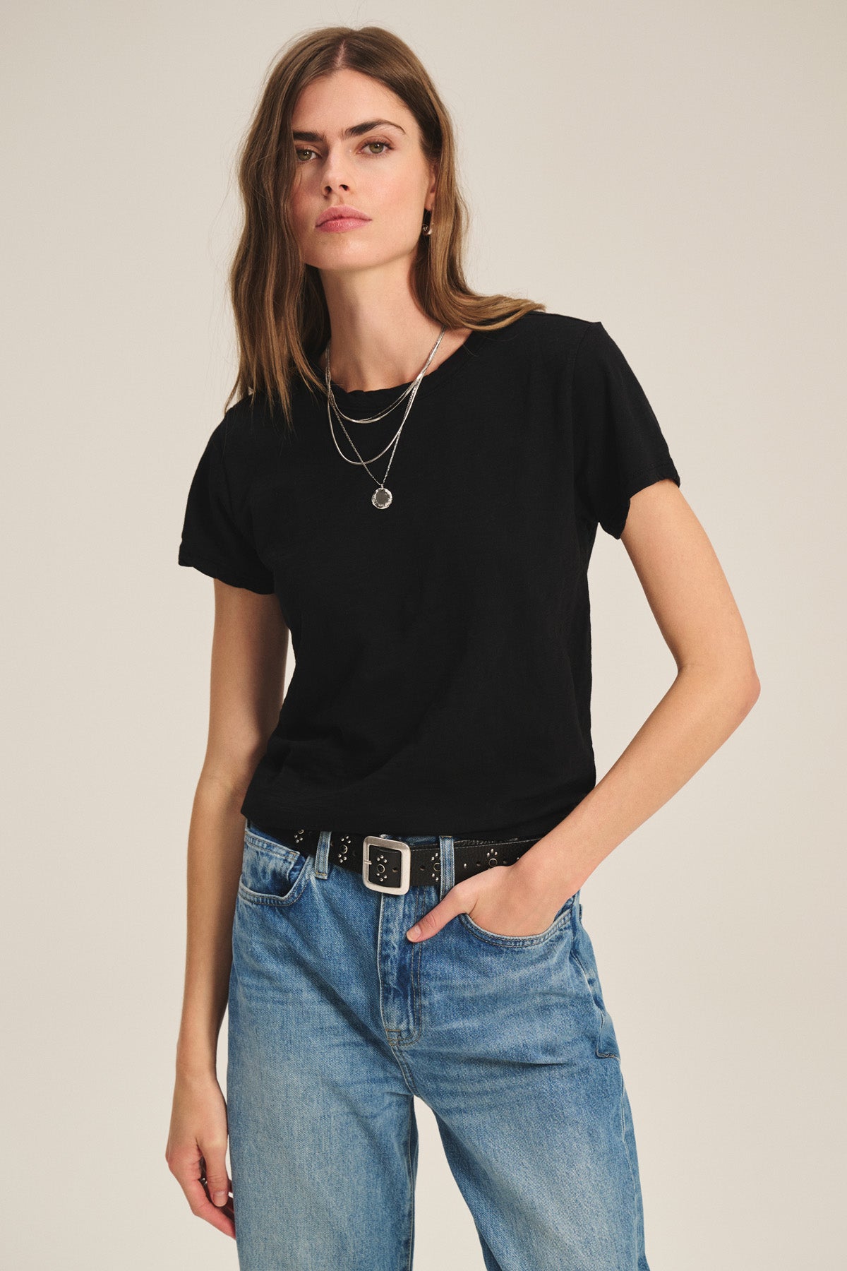 A woman stands against a plain background wearing a black MARIKA TEE by Velvet by Graham & Spencer in a relaxed boxy fit, layered necklaces, blue jeans, and a black belt with a square buckle.