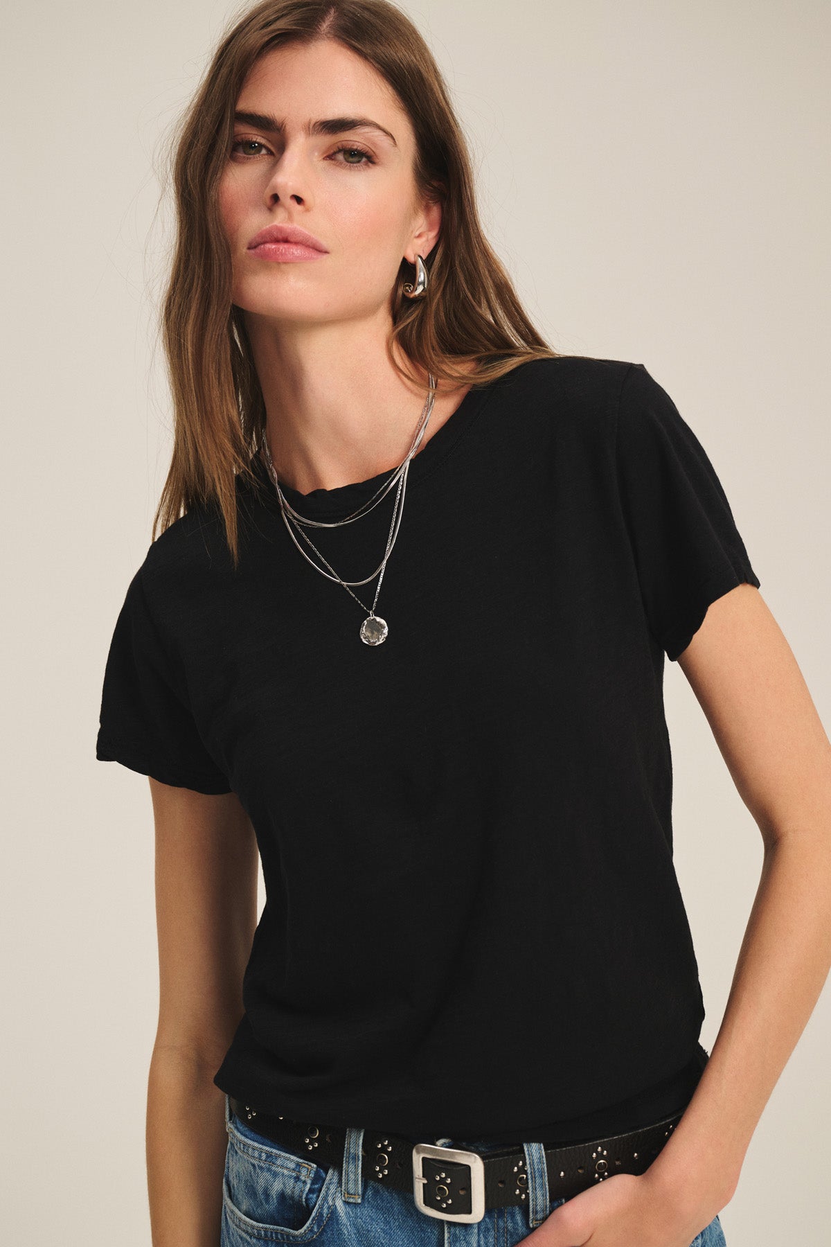 A woman with long brown hair wears the Velvet by Graham & Spencer MARIKA TEE in a relaxed boxy fit, paired with layered silver necklaces and blue jeans with a black belt, standing against a plain background.