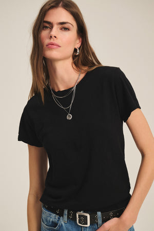 A woman with long brown hair wears the Velvet by Graham & Spencer MARIKA TEE in a relaxed boxy fit, paired with layered silver necklaces and blue jeans with a black belt, standing against a plain background.
