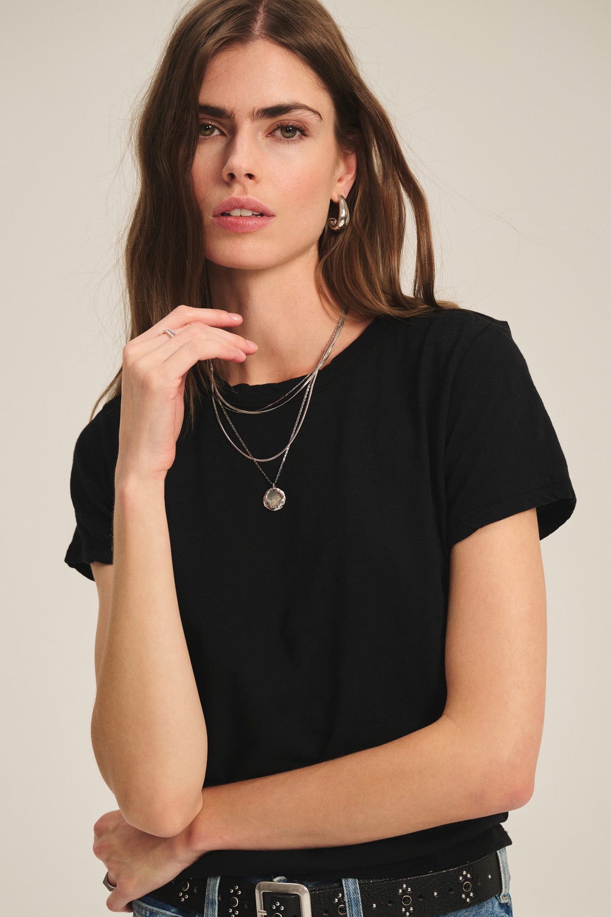 A woman with long brown hair wears a black MARIKA TEE by Velvet by Graham & Spencer in a relaxed, boxy fit, styled with silver necklaces and a studded belt, posing against a plain background.