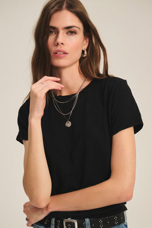 A woman with long brown hair wears a black MARIKA TEE by Velvet by Graham & Spencer in a relaxed, boxy fit, styled with silver necklaces and a studded belt, posing against a plain background.