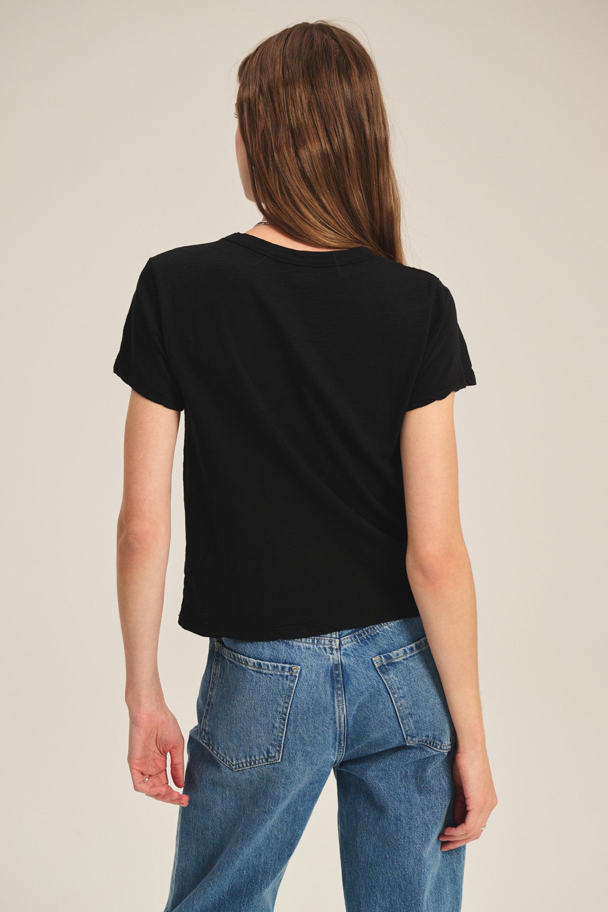 A woman with long brown hair is seen from the back, wearing Velvet by Graham & Spencer's MARIKA TEE—a black cotton slub t-shirt with a relaxed, boxy fit—and blue jeans against a neutral background.
