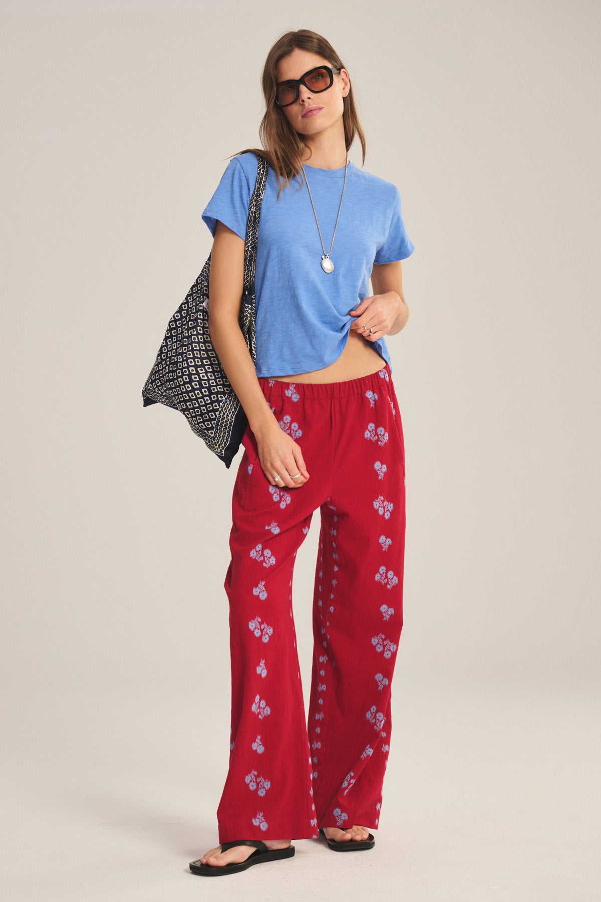 A woman in sunglasses, a blue t-shirt, Velvet by Graham & Spencer’s NELLIE PANT (wide-leg red floral with elastic waist), black sandals, and a black patterned tote bag stands against a plain background.