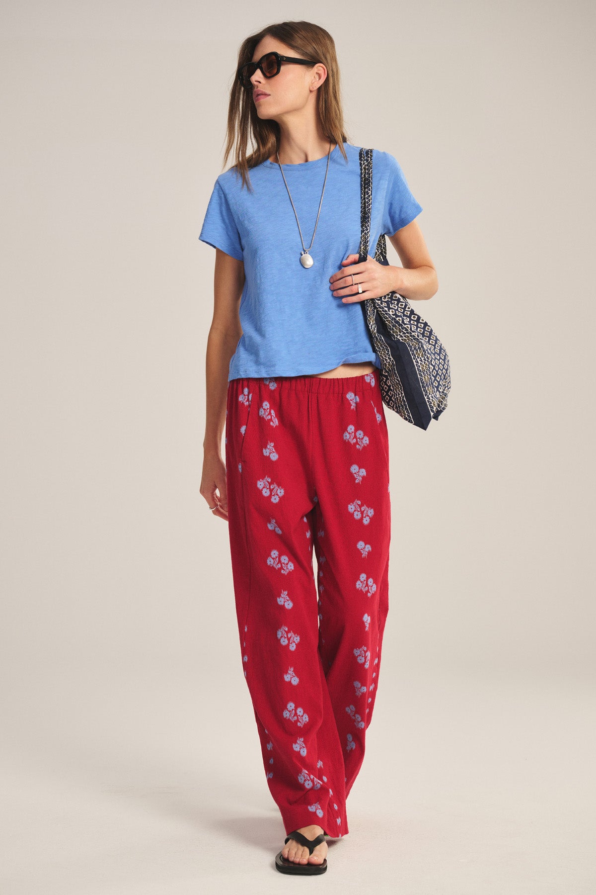 Woman wears a relaxed, boxy-fit blue MARIKA TEE by Velvet by Graham & Spencer in soft cotton slub, red patterned pants, black flip-flops, sunglasses, a necklace, and carries a patterned shoulder bag against a plain background.-41089023738049