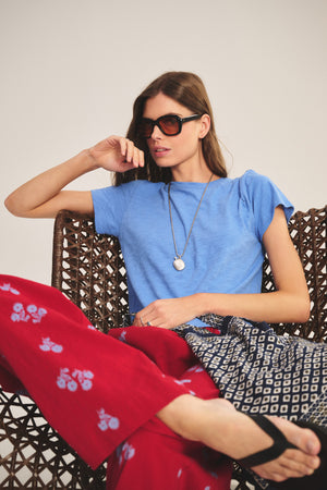 A woman wearing sunglasses, the Velvet by Graham & Spencer MARIKA TEE—a blue cotton slub crew-neck shirt—and patterned red and blue pants sits on a woven chair, one arm on the back and one foot crossed over the other.