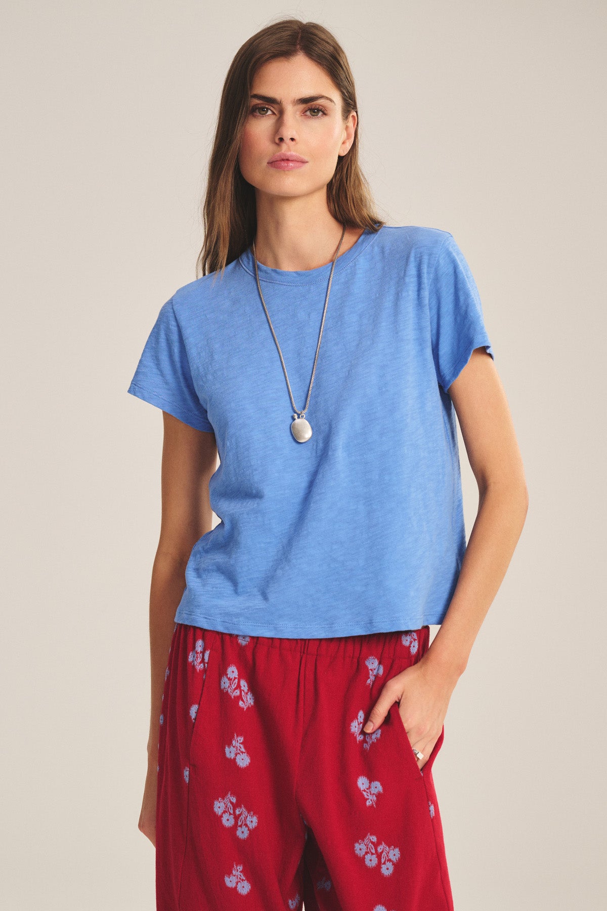 A woman with long hair and a pendant necklace wears the Velvet by Graham & Spencer MARIKA TEE in blue cotton slub, a relaxed boxy fit, paired with red floral pants, standing with one hand in her pocket.-41089023672513