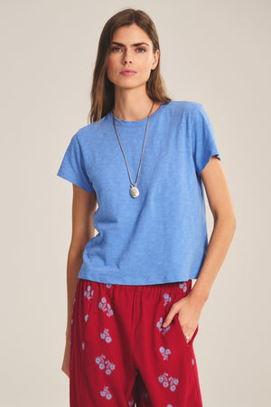 A woman with long hair and a pendant necklace wears the Velvet by Graham & Spencer MARIKA TEE in blue cotton slub, a relaxed boxy fit, paired with red floral pants, standing with one hand in her pocket.