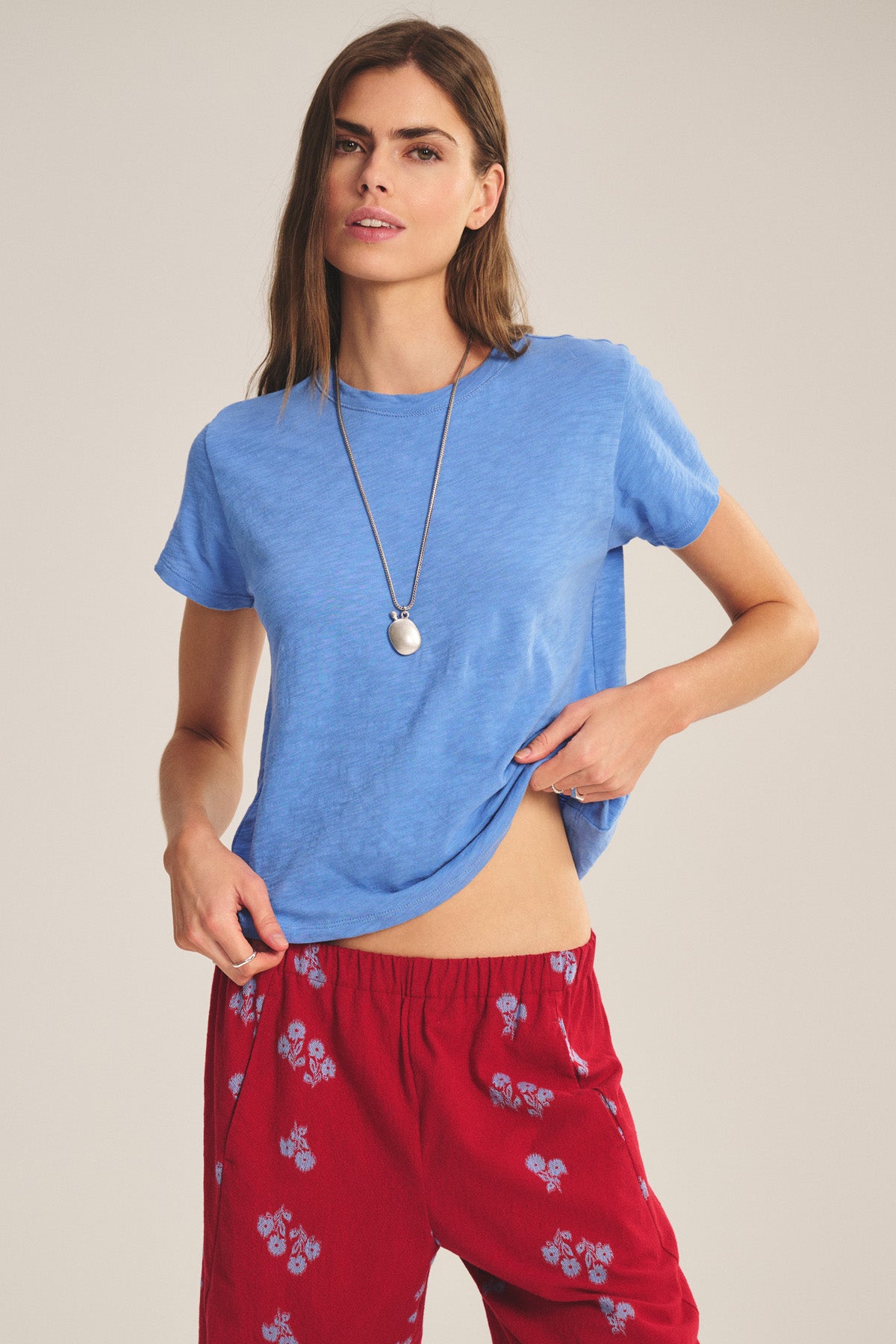 Someone wearing a light blue Velvet by Graham & Spencer MARIKA TEE with a relaxed boxy fit, red floral pants, and a necklace stands against a plain background, holding up one side of the shirt slightly.-41089023639745