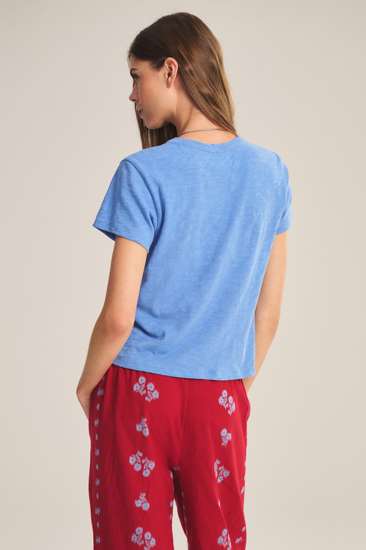 A woman faces away, wearing a light blue MARIKA TEE by Velvet by Graham & Spencer in cotton slub with a relaxed boxy fit, paired with red pants featuring white floral patterns, against a plain background.-41089023606977