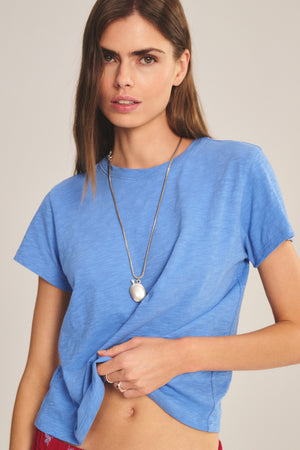 A woman stands against a plain background, holding the hem of her shirt with one hand as she wears the MARIKA TEE by Velvet by Graham & Spencer, featuring a relaxed boxy fit and soft cotton slub fabric. She finishes her effortless look with a long necklace.