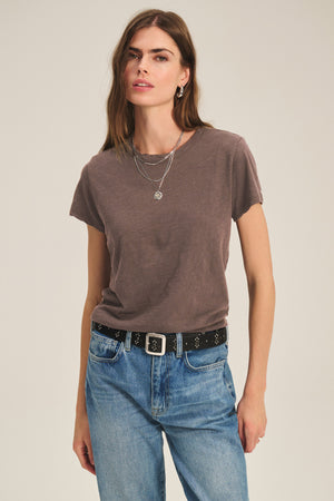 A woman stands against a plain background wearing the brown MARIKA TEE from Velvet by Graham & Spencer in cotton slub with a relaxed boxy fit, blue jeans, a black belt, layered necklaces, and earrings.