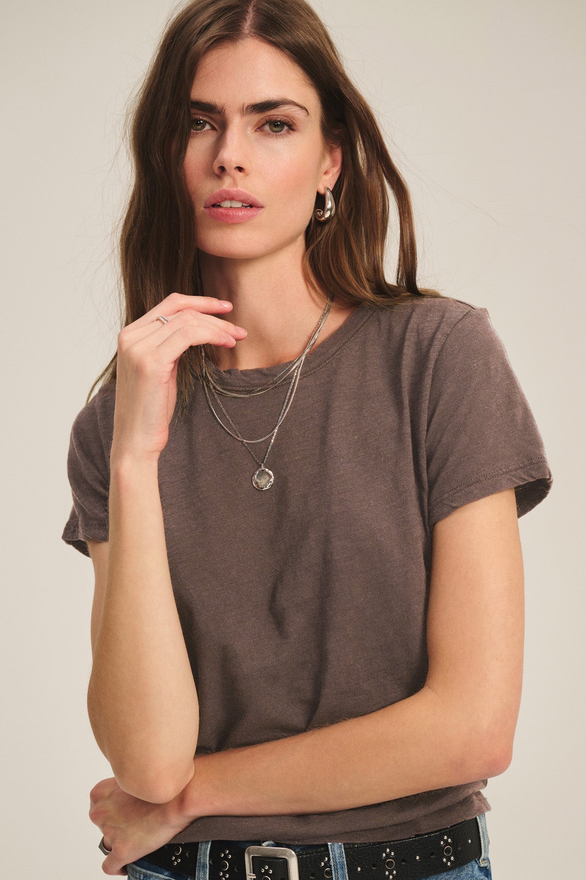 Woman with long brown hair wears the MARIKA TEE by Velvet by Graham & Spencer—a relaxed, boxy-fit cotton slub—styled with layered necklaces and a black belt, posing with hand near face against a plain background.-40921281036481