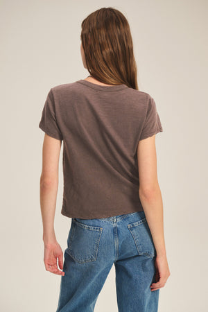 A person with long brown hair faces away from the camera against a plain background, wearing a relaxed boxy fit MARIKA TEE in brown by Velvet by Graham & Spencer and blue jeans.
