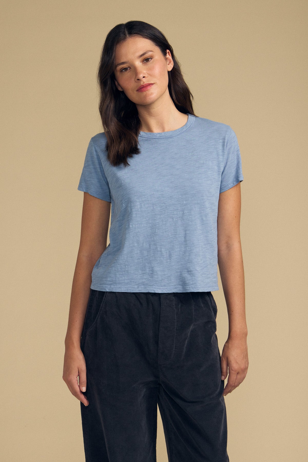 A woman stands against a plain beige background wearing the MARIKA TEE by Velvet by Graham & Spencer—a light blue, boxy-fit tee crafted from soft cotton slub—paired with loose dark pants.