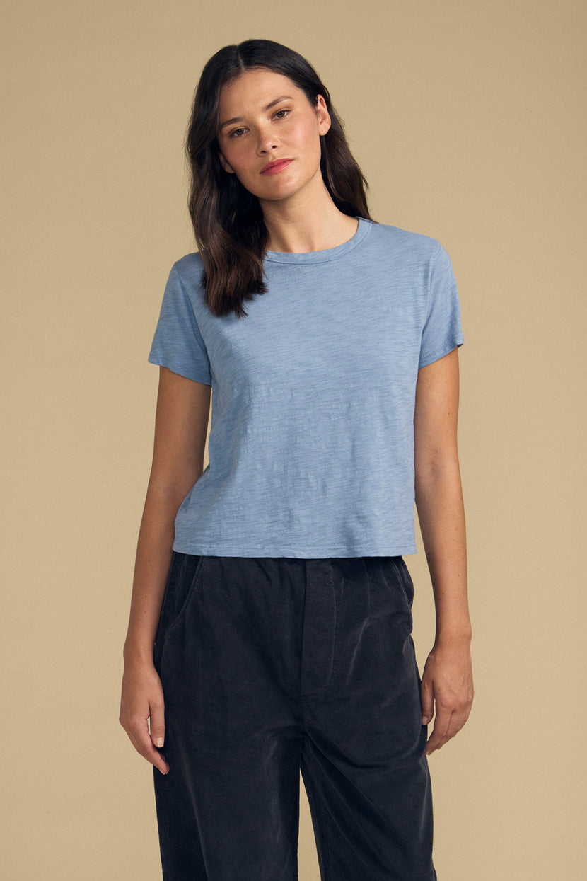 A woman stands against a plain beige background wearing the MARIKA TEE by Velvet by Graham & Spencer—a light blue, boxy-fit tee crafted from soft cotton slub—paired with loose dark pants.