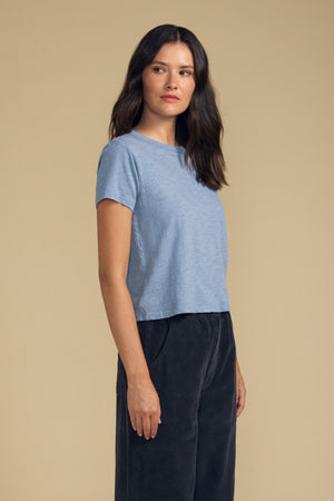 A woman stands against a beige background, slightly turned to the side, wearing the Velvet by Graham & Spencer MARIKA TEE—a light blue cotton slub top with a relaxed boxy fit—paired with dark pants.