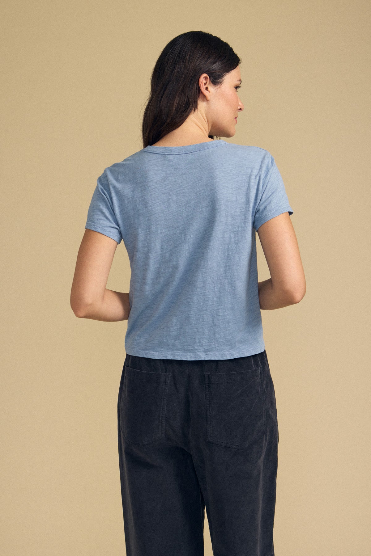 A person with long brown hair is standing with their back to the camera, wearing the Velvet by Graham & Spencer MARIKA TEE in light blue with a boxy fit and dark pants against a beige background.