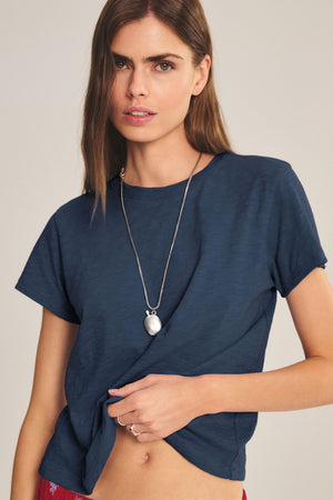 A woman with long brown hair wears a Velvet by Graham & Spencer MARIKA TEE in dark blue cotton slub with a relaxed boxy fit, styled with a silver pendant necklace and posing against a neutral backdrop.