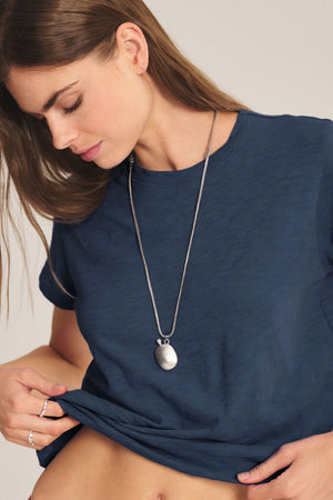 A woman in a blue MARIKA TEE by Velvet by Graham & Spencer, featuring a relaxed boxy fit and cotton slub texture, looks down while slightly lifting the hem of her shirt and accessorizing with a long round-pendant necklace.