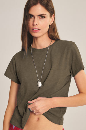 Woman wearing the MARIKA TEE in olive green by Velvet by Graham & Spencer, featuring a cotton slub texture and boxy fit, paired with a long necklace and holding the shirt hem against a plain background.