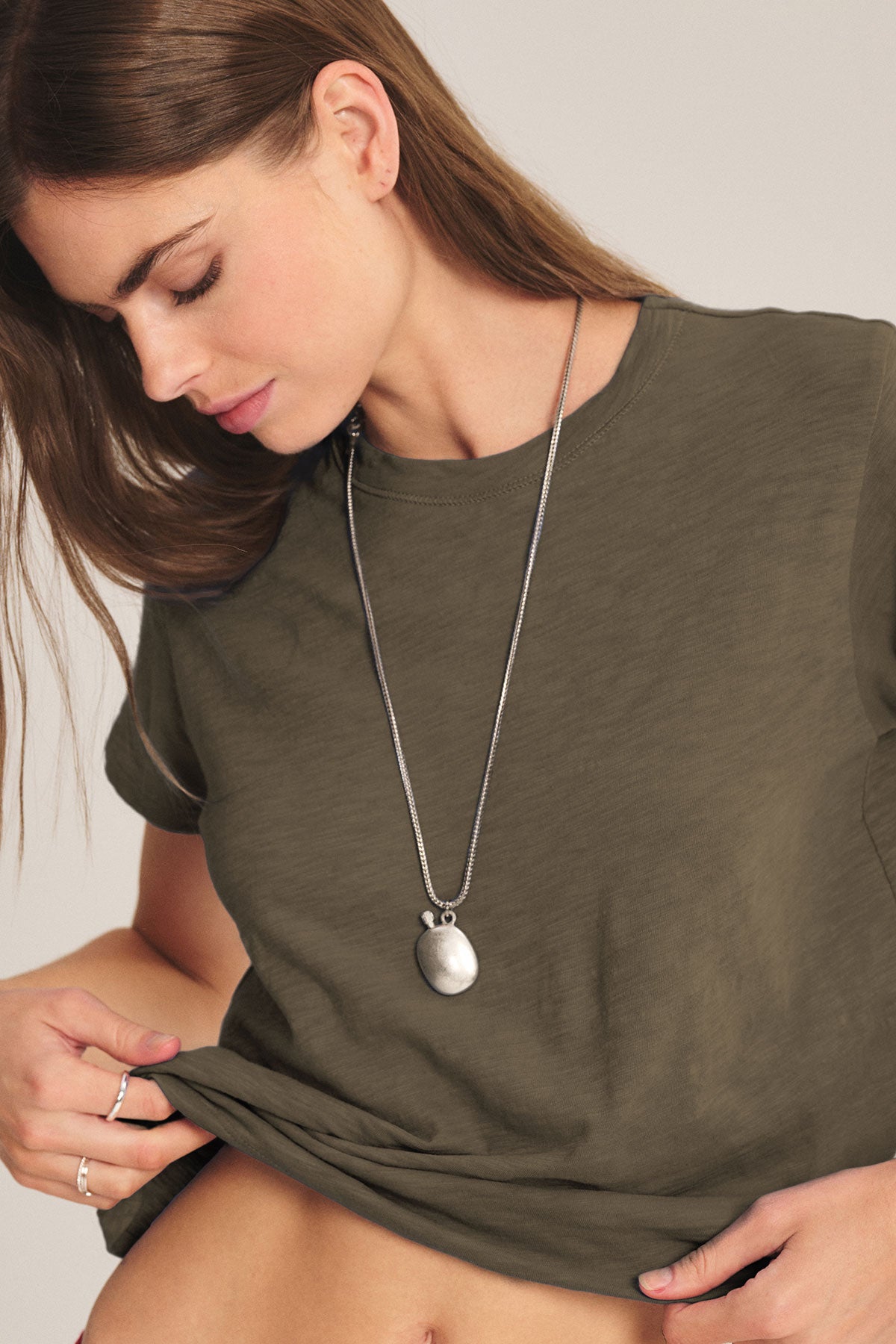 A woman in a boxy-fit olive green cotton slub MARIKA TEE by Velvet by Graham & Spencer and a long necklace looks down while slightly lifting her shirt hem.-40417104658625