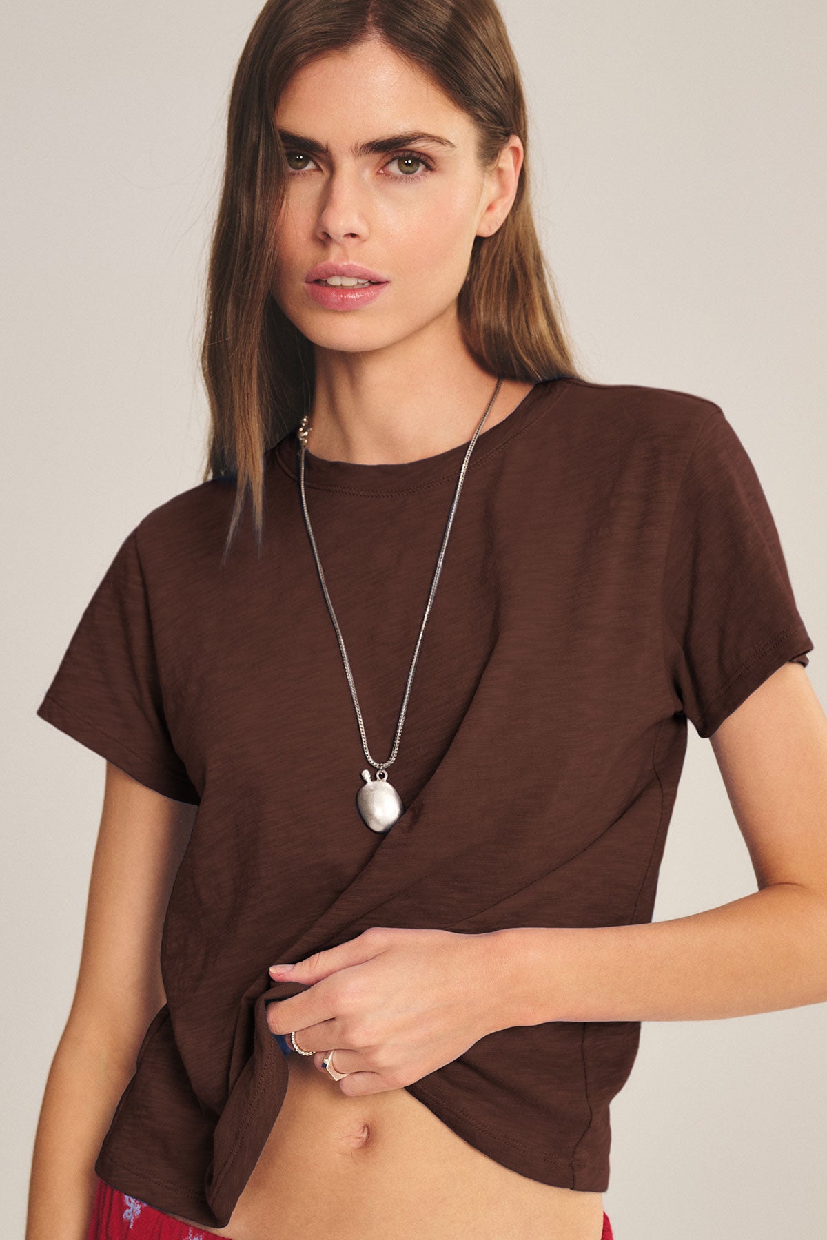A woman with long brown hair wears a brown MARIKA TEE by Velvet by Graham & Spencer in soft cotton slub with a boxy fit, styled with a silver necklace and gently pulling the shirt’s hem.