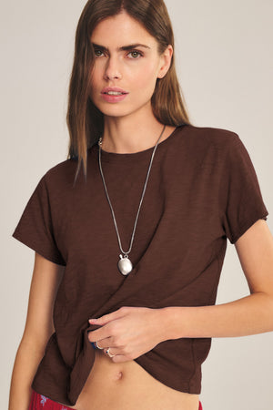 A woman with long brown hair wears a brown MARIKA TEE by Velvet by Graham & Spencer in soft cotton slub with a boxy fit, styled with a silver necklace and gently pulling the shirt’s hem.