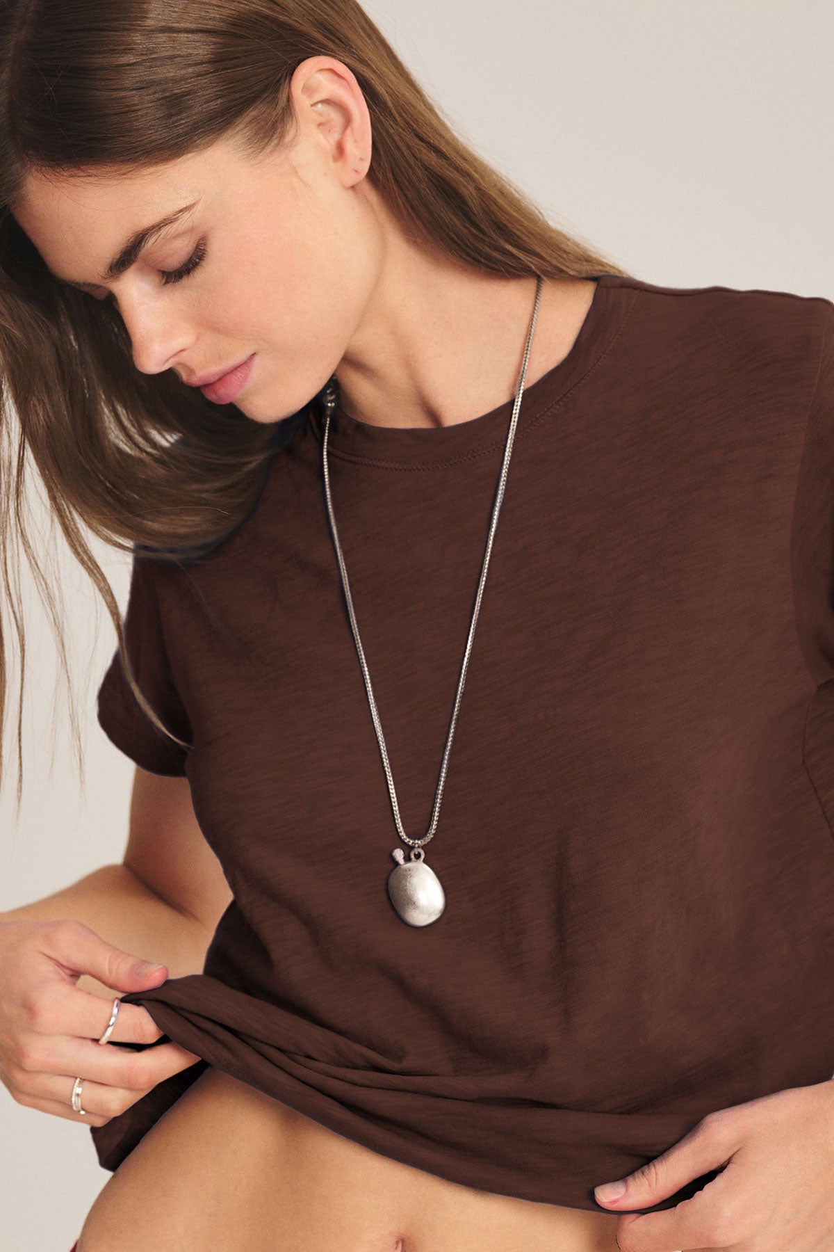 A woman wearing a brown, boxy fit MARIKA TEE by Velvet by Graham & Spencer and a silver pendant necklace looks down as she slightly lifts the hem of her shirt.-40417104822465