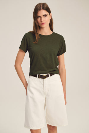 A woman wears the MARIKA TEE by Velvet by Graham & Spencer in dark green, featuring a relaxed boxy fit, tucked into high-waisted, knee-length white shorts with a dark belt.