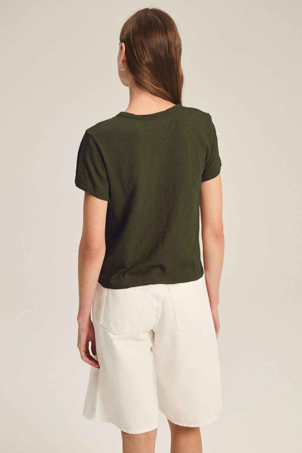 A person with long brown hair stands facing away, wearing Velvet by Graham & Spencer's MARIKA TEE in dark green with a relaxed boxy fit, paired with white knee-length cotton slub shorts, set against a neutral background.