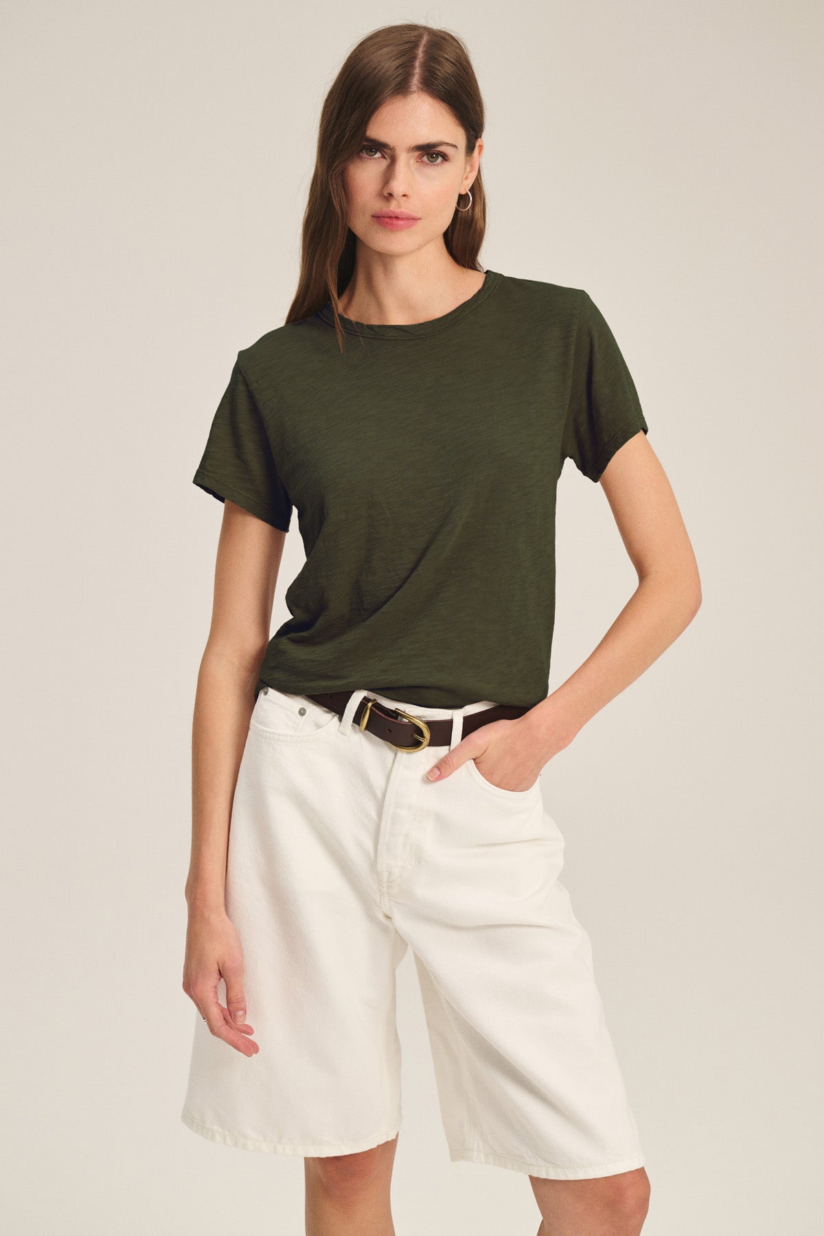 A woman wears a dark green MARIKA TEE by Velvet by Graham & Spencer in a relaxed boxy fit, tucked into high-waisted white shorts with a brown belt, posing with one hand in her pocket against a plain background.
