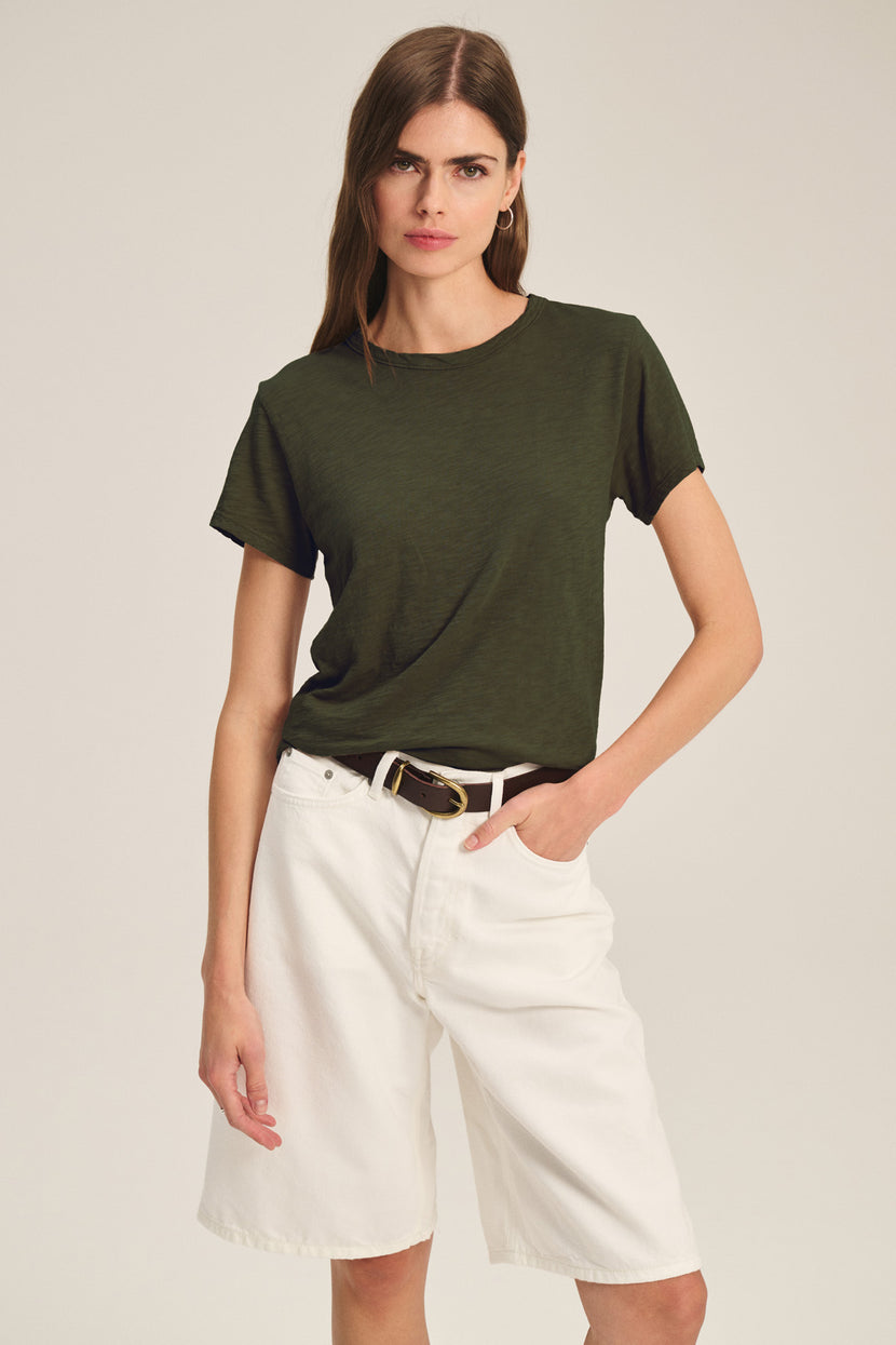 A woman wears a dark green MARIKA TEE by Velvet by Graham & Spencer in a relaxed boxy fit, tucked into high-waisted white shorts with a brown belt, posing with one hand in her pocket against a plain background.