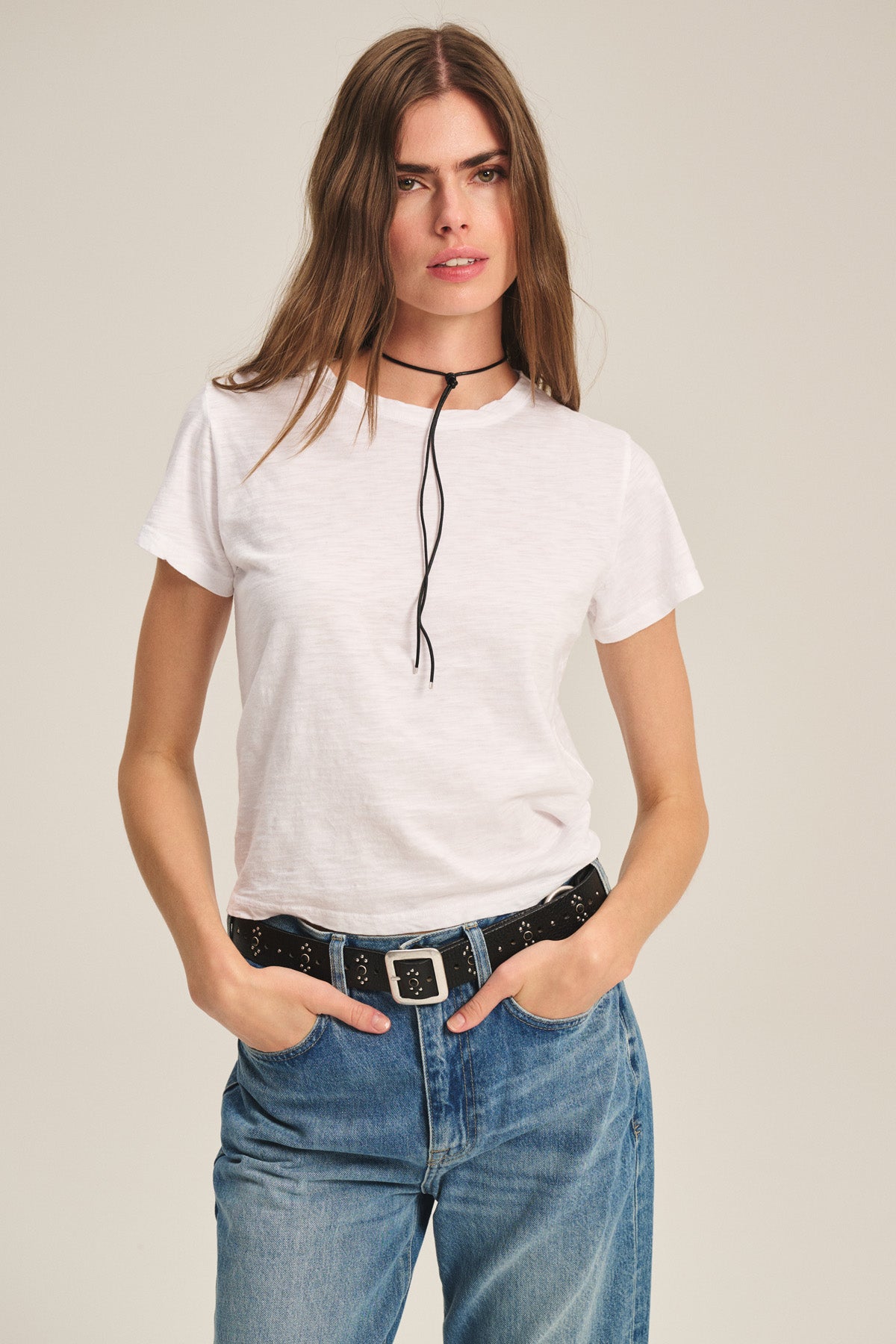 A woman stands with her hands in her jean pockets, wearing the relaxed boxy fit MARIKA TEE by Velvet by Graham & Spencer in soft cotton slub, styled with a black belt, blue jeans, and a thin black choker necklace.
