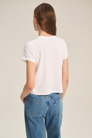 A woman faces away in a white Velvet by Graham & Spencer MARIKA TEE, featuring a cotton slub fabric and relaxed boxy fit, paired with blue jeans against a plain background.