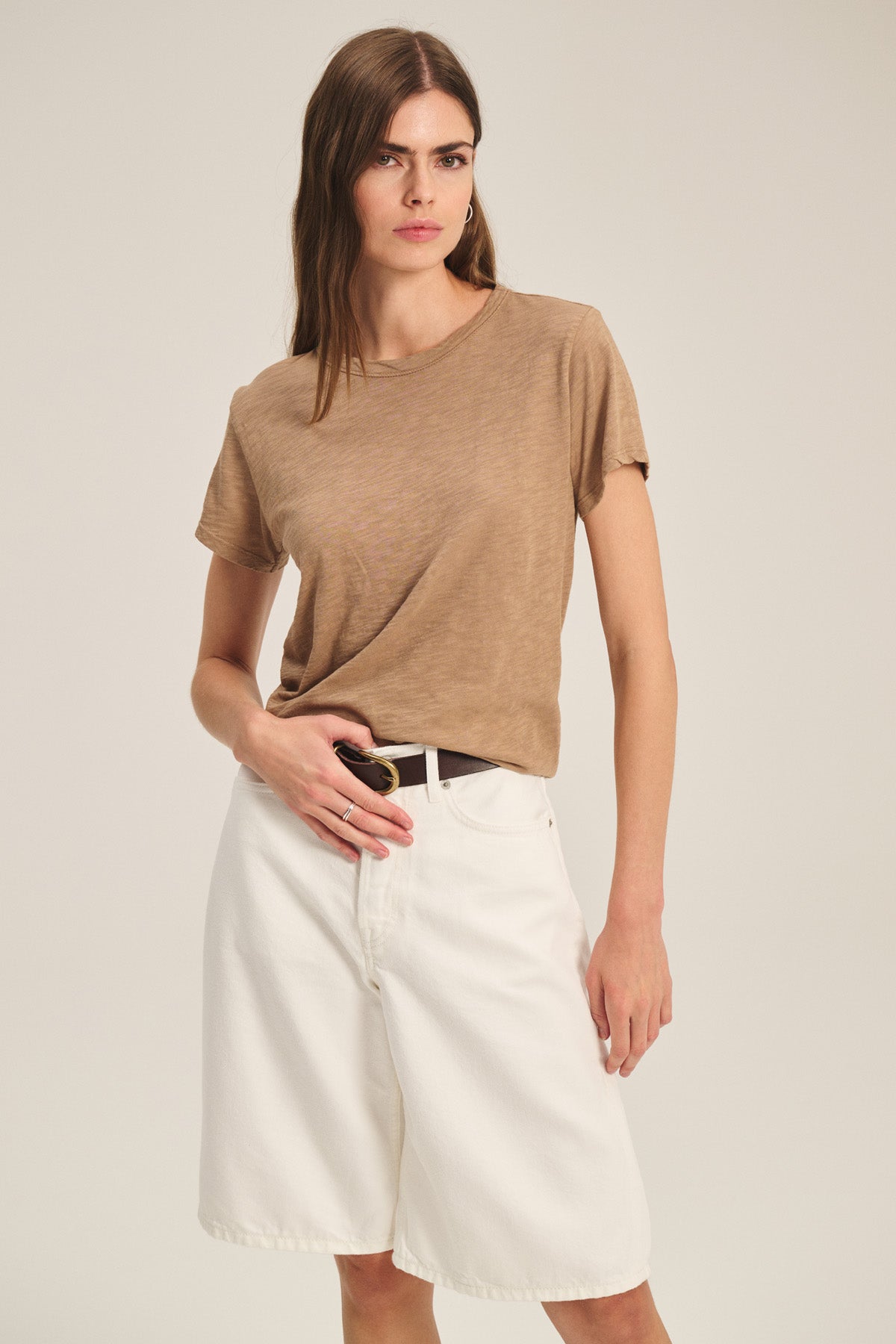 Wearing the MARIKA TEE by Velvet by Graham & Spencer—a brown cotton slub short-sleeve t-shirt with a classic crew neckline—tucked into high-waisted white knee-length shorts, this look exudes minimalist style. The relaxed, boxy fit of the tee adds an effortless touch against the plain, light background.