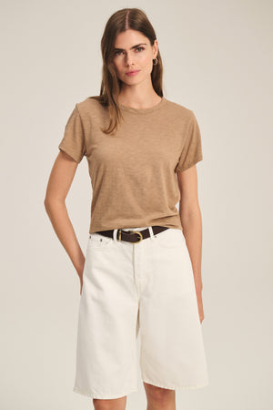 A woman stands facing forward in a light brown MARIKA TEE by Velvet by Graham & Spencer, styled with white knee-length shorts and a black belt against a neutral background.