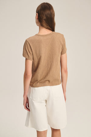A person with long brown hair, wearing a boxy-fit brown MARIKA TEE by Velvet by Graham & Spencer and white knee-length shorts, stands facing away from the camera against a plain background.