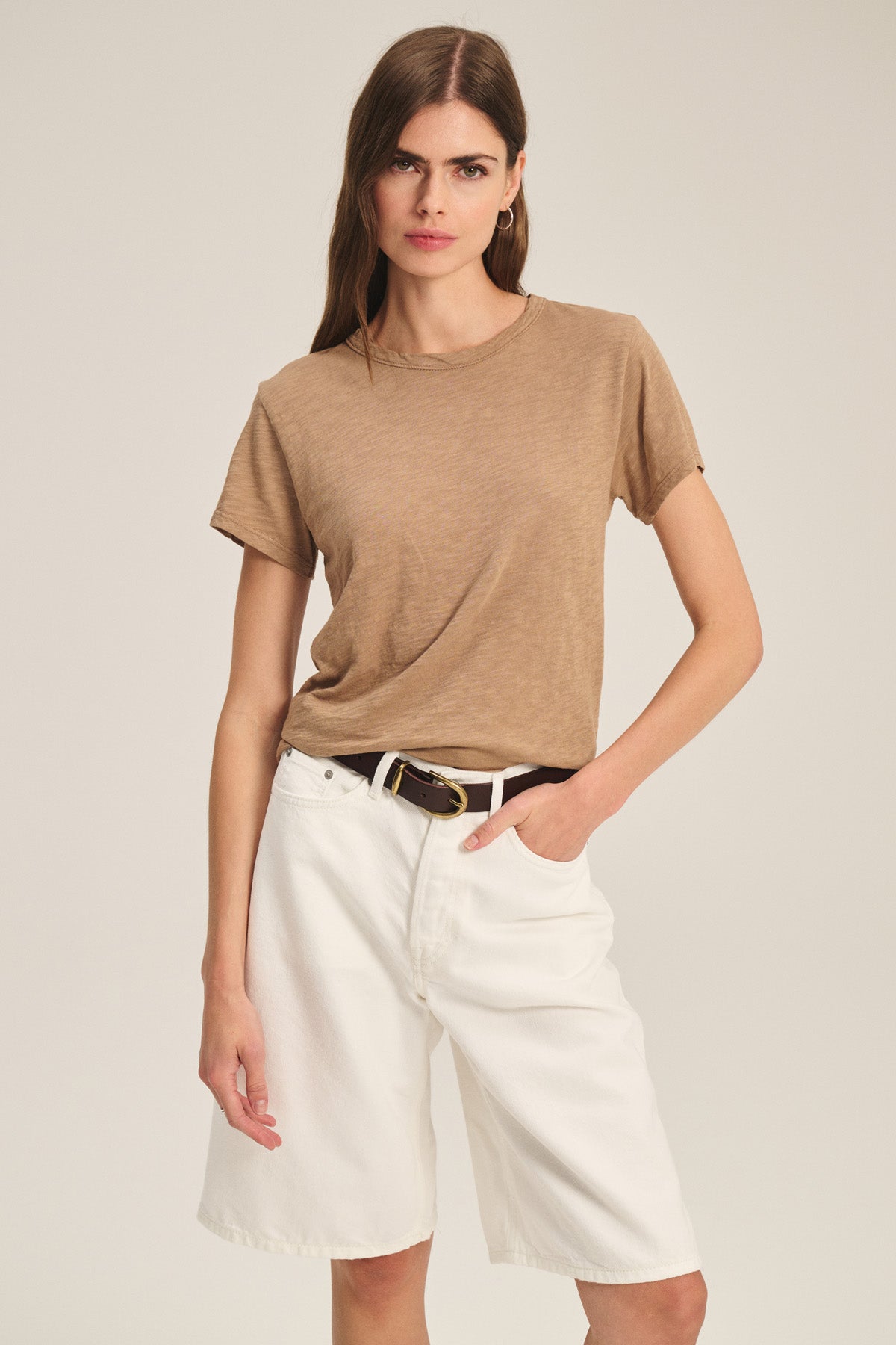 A woman faces forward, wearing the MARIKA TEE by Velvet by Graham & Spencer in brown cotton slub with a boxy fit, tucked into white knee-length shorts with a black belt. One hand is in her pocket against a plain background.