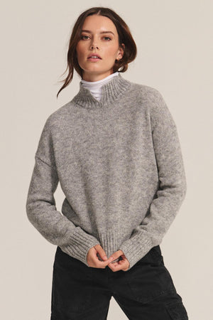 A woman stands against a plain background wearing the BRIELLE SWEATER by Velvet by Graham & Spencer over a white turtleneck and black pants, holding the sweater’s hem.